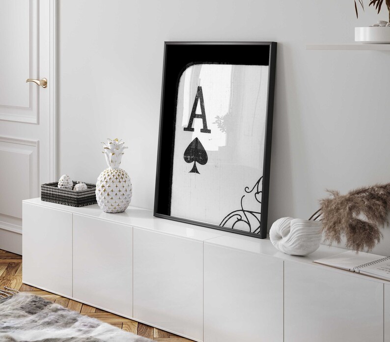 Op de afbeelding: Een zwart ingelijst afdruk van de Aas van Schoppen met een witte achtergrond. De afdruk hangt aan een witte muur boven een wit kastje met drie lades. Er staat een witte ananas en een witte schelp op het kastje.