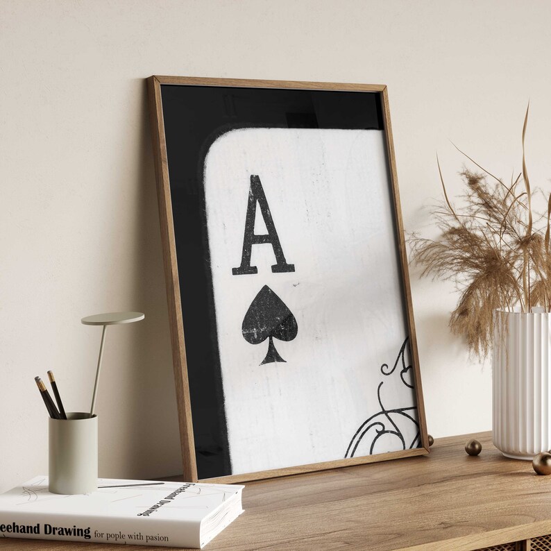 Op de afbeelding: Een ingelijste print van een speelkaart, de Aas van Schoppen, met een zwart-wit ontwerp. De kaart is op een witte achtergrond met een zwarte rand.