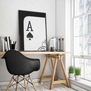 Op de afbeelding: Een zwart-witte ingelijste print van de Aas van Schoppen met een decoratieve krul in de hoek. De print hangt aan een witte muur boven een houten bureau met een zwarte stoel en een bril.
