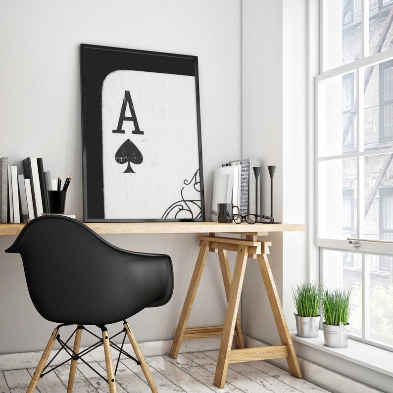 Op de afbeelding: Een zwart-witte ingelijste print van de Aas van Schoppen met een decoratieve krul in de hoek. De print hangt aan een witte muur boven een houten bureau met een zwarte stoel en een bril.