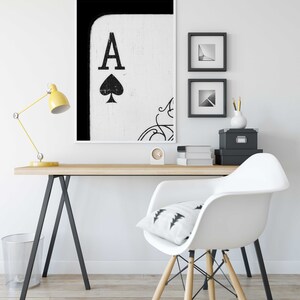 Op de afbeelding: Een zwart-witte print van de Aas van Schoppen speelkaart in een wit frame hangend aan een witte muur boven een houten bureau met een witte stoel en een gele lamp.