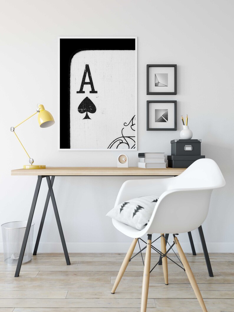 Op de afbeelding: Een zwart-witte print van de Aas van Schoppen speelkaart in een wit frame hangend aan een witte muur boven een houten bureau met een witte stoel en een gele lamp.
