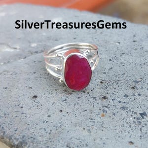May include: A silver ring with a bright red ruby gemstone set in a simple oval shape. The ring has three bands that wrap around the finger.