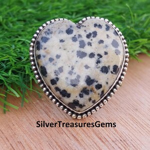 May include: A heart-shaped gemstone ring with a speckled cream and black pattern. The stone is set in a silver-coloured metal band with a beaded edge. The text "SilverTreasuresGems" is at the bottom.