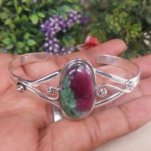 May include: A silver cuff bracelet with an oval gemstone in shades of red and green. The bracelet has a decorative design with swirling details. The gemstone is set in a silver frame.