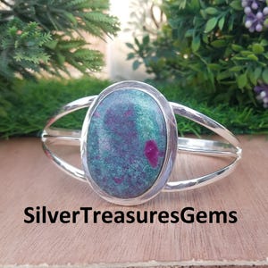 May include: A silver cuff bracelet featuring an oval gemstone. The gemstone displays a blend of green and purple shades with red flecks. The text "SilverTreasuresGems" is visible at the bottom of the image.
