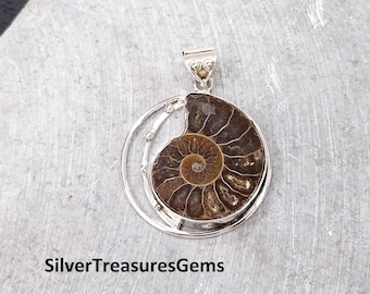 May include: Silver pendant featuring a fossilized ammonite in a circular setting. The ammonite is brown and has a spiral pattern. The pendant is on a gray background.