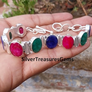 May include: A silver bracelet featuring oval-cut gemstones in various colors: red, green, and blue. The bracelet has a chain link design and the text "SilverTreasuresGems".