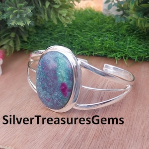 Puede incluir: Una pulsera de plata con una piedra preciosa ovalada en tonos verdes y morados. La piedra preciosa está engastada en un bisel de plata, y la pulsera tiene un diseño de caña dividida. El texto "SilverTreasuresGems" está en la parte inferior.