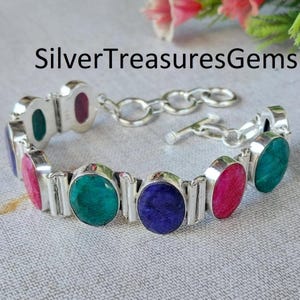 May include: Silver bracelet featuring oval-shaped gemstones in shades of green, pink, and blue. The bracelet has a silver link chain and a toggle clasp. The text "SilverTreasuresGems" is visible in the image.