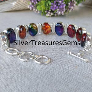 May include: A silver bracelet featuring oval-shaped gemstones in various colors, including purple, red, orange, and green. The bracelet has a toggle clasp and the text "SilverTreasuresGems" is visible.