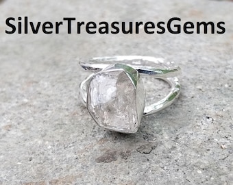 May include: A silver ring with a large, clear, irregularly shaped stone. The ring has a double band design. The text "SilverTreasuresGems" is at the top of the image. The ring is set on a gray stone surface.