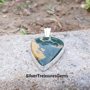 Puede incluir: Un colgante de plata con una piedra verde y amarilla en forma de lágrima. La piedra tiene un patrón único de remolinos y manchas verdes y amarillas.