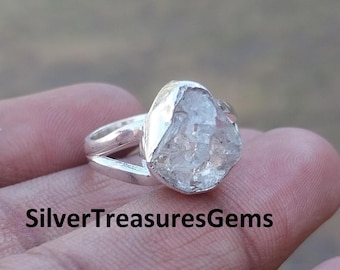 May include: A silver ring with a large, irregularly shaped, clear crystal stone. The ring band splits into two parts on either side of the stone. The text "SilverTreasuresGems" is at the bottom of the image.