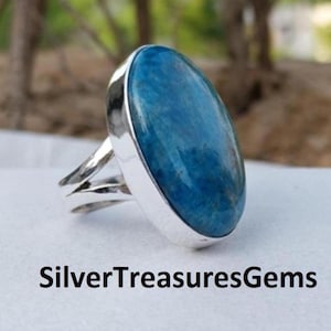 May include: A silver ring with a large, oval-shaped blue gemstone. The gemstone has a smooth, polished surface. The ring's band splits into two parts on each side of the gemstone. The text "SilverTreasuresGems" is at the bottom of the image.