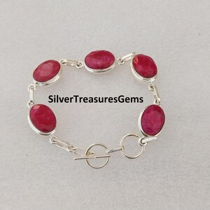 Puede incluir: Una pulsera de cadena plateada con cuatro piedras preciosas ovaladas de color rojo rosado intenso engastadas en biseles de metal liso. La pulsera incluye un cierre de barra y lleva el texto SilverTreasuresGems en el centro del diseño.