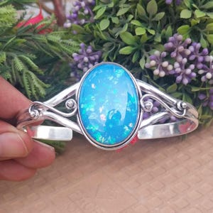 Peut inclure: Un bracelet manchette en argent avec une pierre d'opale ovale bleu clair. La pierre a un aspect chatoyant et irisé. Le bracelet présente des volutes décoratives. Fond de feuillage vert et violet.
