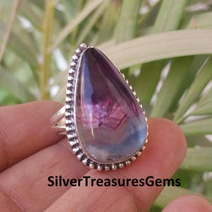Può includere: Anello d'argento con una grande pietra a forma di goccia. La pietra presenta sfumature di viola, rosa e blu. L'anello ha un bordo perlato e il testo "SilverTreasuresGems" è visibile.
