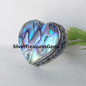 May include: Heart-shaped abalone shell pendant with iridescent blues and greens, set in a silver-toned metal frame. The frame features an ornate, detailed design. The text "SilverTreasuresGems" is visible on the pendant.