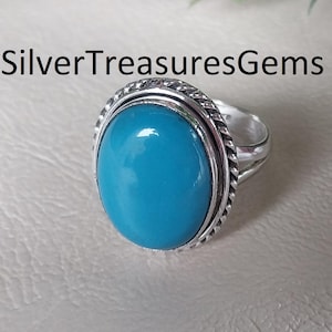 May include: A silver ring with a large, oval, bright turquoise stone. The stone is set in a silver band with a twisted rope design around the stone. The text "SilverTreasuresGems" is above the ring.