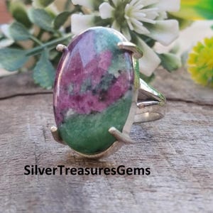 May include: Oval-shaped ring featuring a large, polished gemstone with a mix of green and magenta hues. The stone is set in a silver band with decorative prongs. The text "SilverTreasuresGems" is at the bottom.