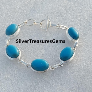 May include: A silver chain bracelet featuring four oval-shaped, smooth turquoise-colored stones set in silver bezels. The bracelet includes a toggle clasp closure and is marked with the text SilverTreasuresGems in the center of the image.