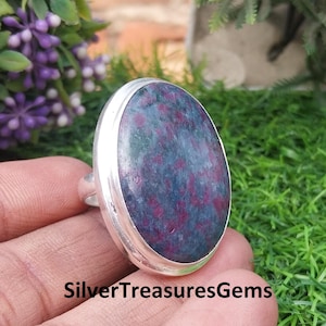 May include: A silver ring featuring a large, oval-shaped gemstone. The gemstone has a dark blue-green base color with red speckles. The ring is held in a hand against a background of green grass and purple flowers. The text "SilverTreasuresGems" is visible.