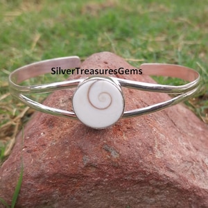May include: A silver cuff bracelet featuring a white and brown spiral design at its centre. The bracelet is displayed on a red rock, with green grass in the background. The text "SilverTreasuresGems" is visible.