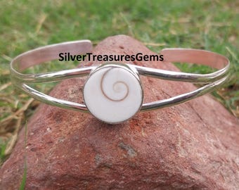 May include: A silver cuff bracelet with a white and brown spiral-shaped centerpiece. The bracelet is resting on a red rock against a backdrop of green grass. The text "SilverTreasuresGems" is visible.
