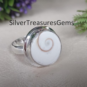 May include: A silver ring with a large, round, white stone with a spiral pattern. The ring has a polished finish and the text "SilverTreasuresGems" is visible.
