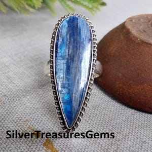 Op de afbeelding: Een opvallende ring met een grote, blauwe kyaniet edelsteen. De steen is gezet in een zilverkleurig metalen frame met een decoratieve, touwachtige rand. De ring wordt gepresenteerd op een neutrale ondergrond. De tekst "SilverTreasuresGems" is zichtbaar.