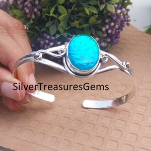 Puede incluir: Una pulsera de plata con una gema ovalada de color azul brillante. La pulsera abierta tiene diseños decorativos y el texto "SilverTreasuresGems" es visible.