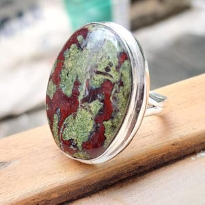May include: Oval-shaped ring featuring a large, polished stone with a mottled pattern of red and green hues. The stone is set in a silver-colored metal band. The ring is displayed on a wooden surface.
