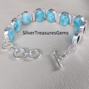 May include: A silver bracelet featuring oval-shaped, light blue gemstones. The bracelet has a toggle clasp and is displayed on a neutral surface. The text "SilverTreasuresGems" is visible on the image.