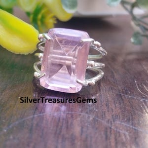 May include: A silver ring with a large, rectangular, light pink gemstone. The ring has a multi-band design with small decorative elements. The text "SilverTreasuresGems" is visible on the surface.