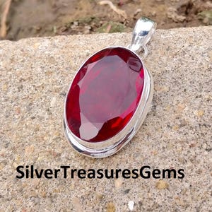 May include: A silver pendant with a large, oval-shaped, deep red gemstone. The gemstone is set in a silver bezel and has a polished surface. The pendant has a silver bail at the top for attaching to a chain. The text "SilverTreasuresGems" is at the bottom.