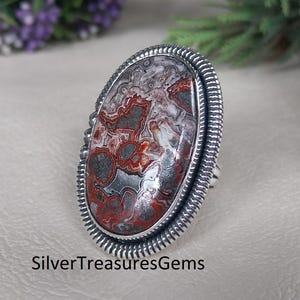 May include: Oval-shaped silver ring featuring a large, patterned stone with red, gray, and white swirls. The stone is set in a textured silver band. The ring is a statement piece, perfect for adding a touch of elegance to any outfit. The text "SilverTreasuresGems" is at the bottom.