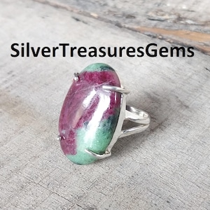 Peut inclure: Bague ovale ornée d'une grosse pierre précieuse polie aux teintes rouge, verte et blanche. La pierre est sertie dans un anneau en métal argenté. Le texte "SilverTreasuresGems" est affiché au-dessus de la bague.