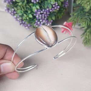 May include: A silver cuff bracelet with a large, oval seashell. The shell is a mix of brown and white tones, set in a simple frame. The bracelet has a minimalist design. The bracelet is a stylish piece of jewellery.