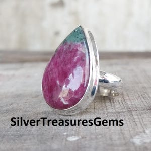 Op de afbeelding: Een zilveren ring met een grote, druppelvormige edelsteen. De edelsteen is voornamelijk diep magenta van kleur, met een groen gedeelte aan de bovenkant. De band en zetting van de ring zijn zilver. De tekst "SilverTreasuresGems" staat onderaan.