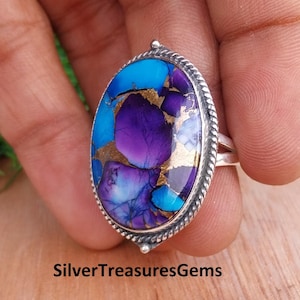 May include: Oval-shaped silver ring featuring a large, vibrant gemstone. The stone showcases a mix of blue, purple, and gold hues. The ring's band is a simple silver design, and the gemstone is set within a detailed silver bezel. The text "SilverTreasuresGems" is at the bottom.