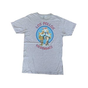 Peut inclure: Un t-shirt gris chiné avec un motif de dessin animé de deux poulets dans un cercle bleu, avec l'inscription "LOS POLLOS HERMANOS" en rouge et bleu. Le t-shirt a un col rond et des manches courtes.