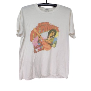 May include: A white short-sleeve t-shirt featuring a graphic print of Jimi Hendrix playing a guitar. The text "Jimi Hendrix" appears in large orange letters above the image, with the phrase "Just ask the ears" printed in smaller orange script below.
