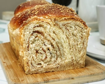 May include: A loaf of freshly baked bread with a golden-brown crust and a swirl pattern, topped with crunchy crumbs. The bread sits on a small wooden cutting board. A white teacup and teapot are in the background.