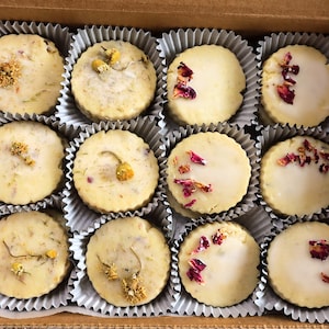 May include: A box of twelve round, light-colored cookies, each nestled in a fluted paper cup. Some cookies are topped with dried flowers, while others have a white glaze and scattered pink petals. The box has floral patterned paper.