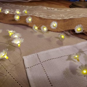 May include: A string of daisy-shaped fairy lights with yellow centres. The lights are strung on a thin wire and are arranged on a table with a burlap runner and white linen cloth. The lights are illuminated.