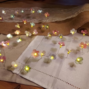 May include: A garland of fairy lights with small, pastel-coloured flower blossoms. The flowers are in shades of pink, white, and blue, each with a warm yellow light. The lights are arranged on a thin, silver wire.