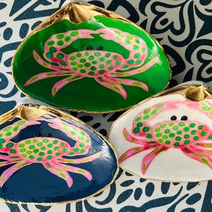 May include: Three painted clam shells with gold rims. Each shell features a crab design in pink and green on a background of green, navy blue, and white. The shells are displayed on a patterned blue and white surface.