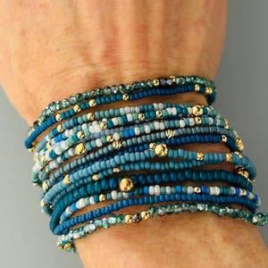 May include: A stack of beaded bracelets in shades of blue, teal, and white, accented with gold-colored beads. The bracelets are worn on a wrist, showcasing the variety of colors and textures.