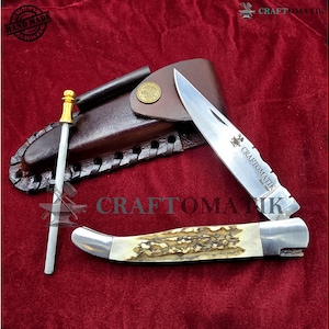 Peut inclure: Un ensemble de couteaux artisanal avec un étui en cuir, une tige d'affûtage et un couteau avec un manche en os. La lame du couteau est gravée de "CRAFTOMATIK". L'ensemble est présenté sur une surface de velours rouge.